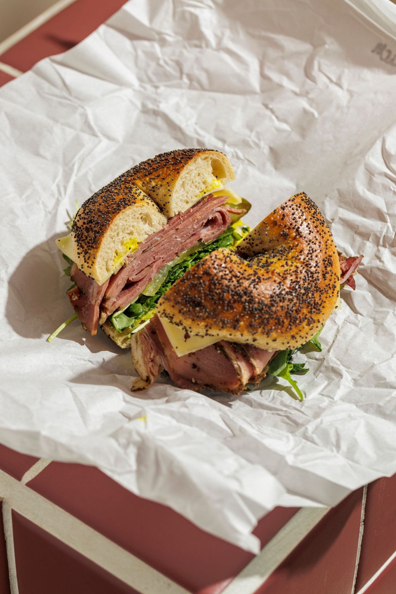 A pastrami Deli Stack bagel resting on white paper wrapping on a burgundy tile.