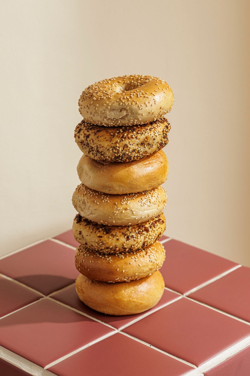A stack of freshly baked bagels — sesame, everything, plain — on a burgundy tile.