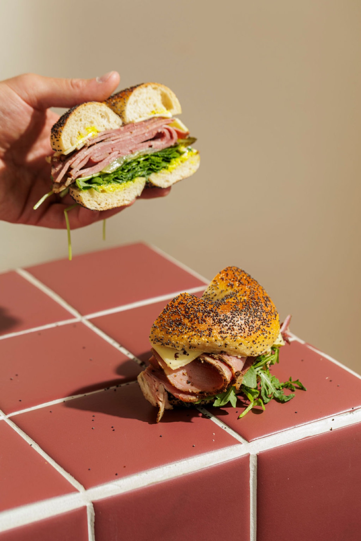 A Deli Stack pastrami bagel — dripping with mustard mayo — held above a burgundy tile plinth next to a second everything-bagel version on the tile.