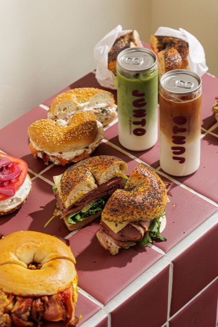A spread of JUNTO bagels — pastrami, salmon and cream cheese — alongside iced matcha and iced coffee cans on a burgundy tiled table.