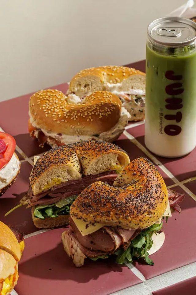 A takeaway coffee cup, pickled onion and cream cheese bagel, pastrami bagel and JUNTO iced drinks arranged on the burgundy tile counter.