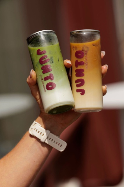 A hand holding two JUNTO cans together — iced matcha and iced coffee — in warm daylight.
